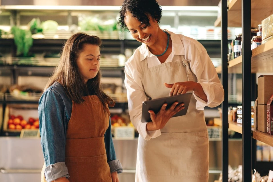 Girl with down syndrome at work in grocery store with carer looking at ipad