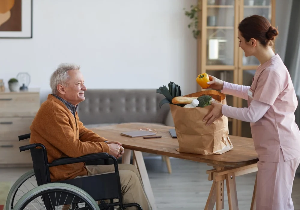 Elderly man in wheelchair smiling at nurse who is unpacking groceries